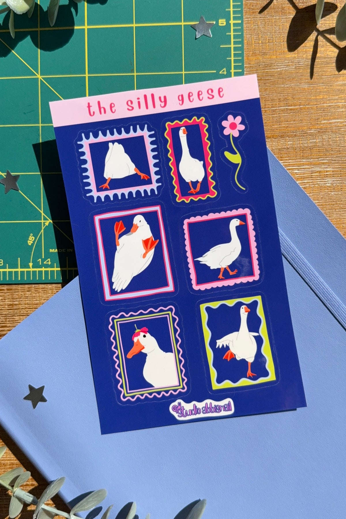 A square sticker sheet titled 'the silly geese' with six illustrated goose stamps on a dark blue background, styled on top of a blue notebook with eucalyptus leaves and a green cutting mat.