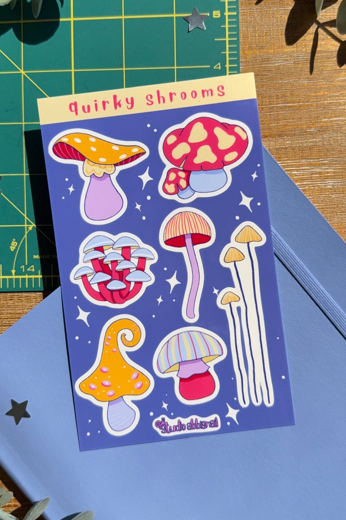 A sticker sheet titled 'Quirky shrooms' featuring colorful mushroom illustrations on a blue background, styled on a wooden surface with a blue notebook, green cutting mat, and eucalyptus leaves.