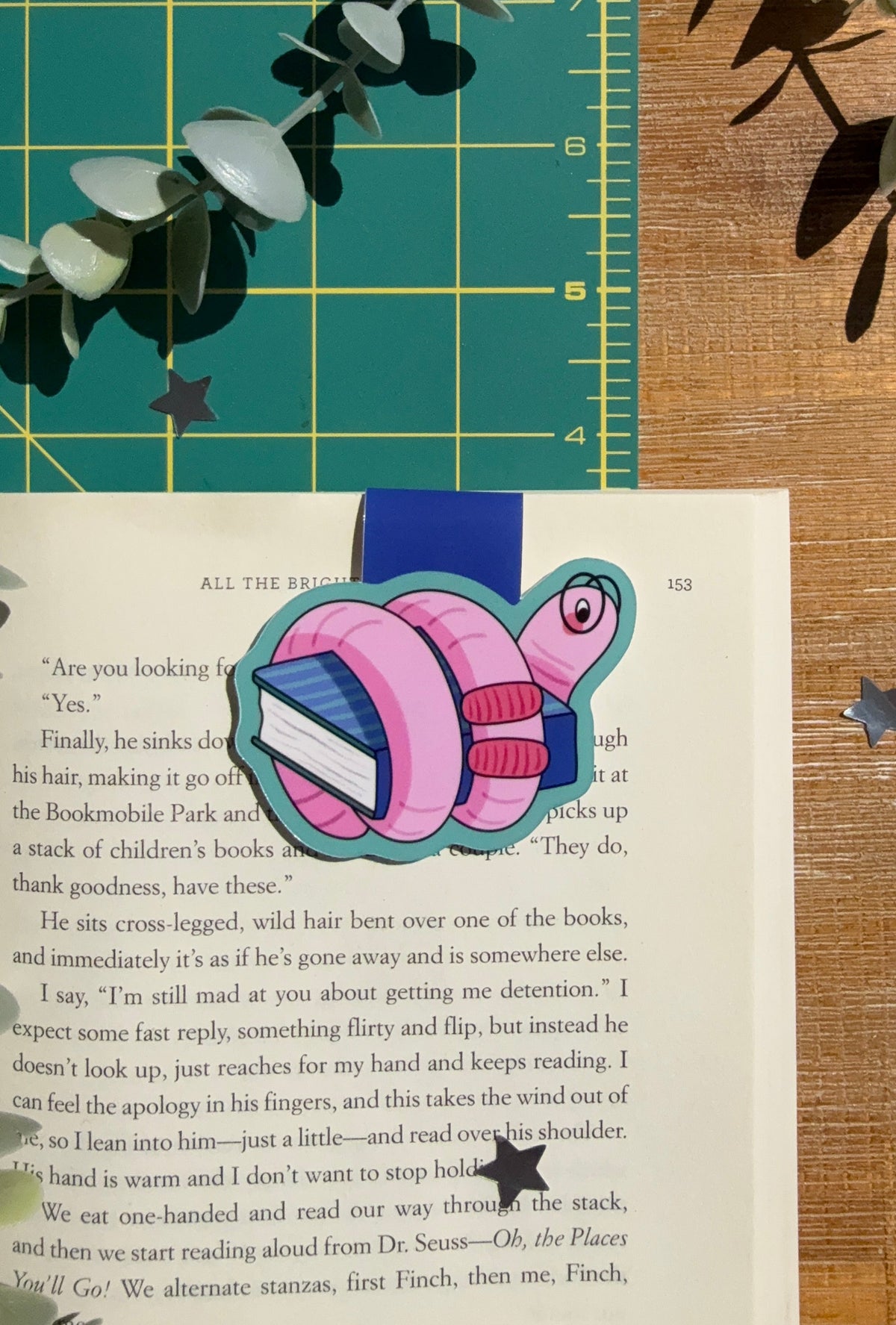 A magnet bookmark of a pink worm with glasses wrapped around a blue book, sitting on an open page book with a green cutting mat and wood desk in the background.
