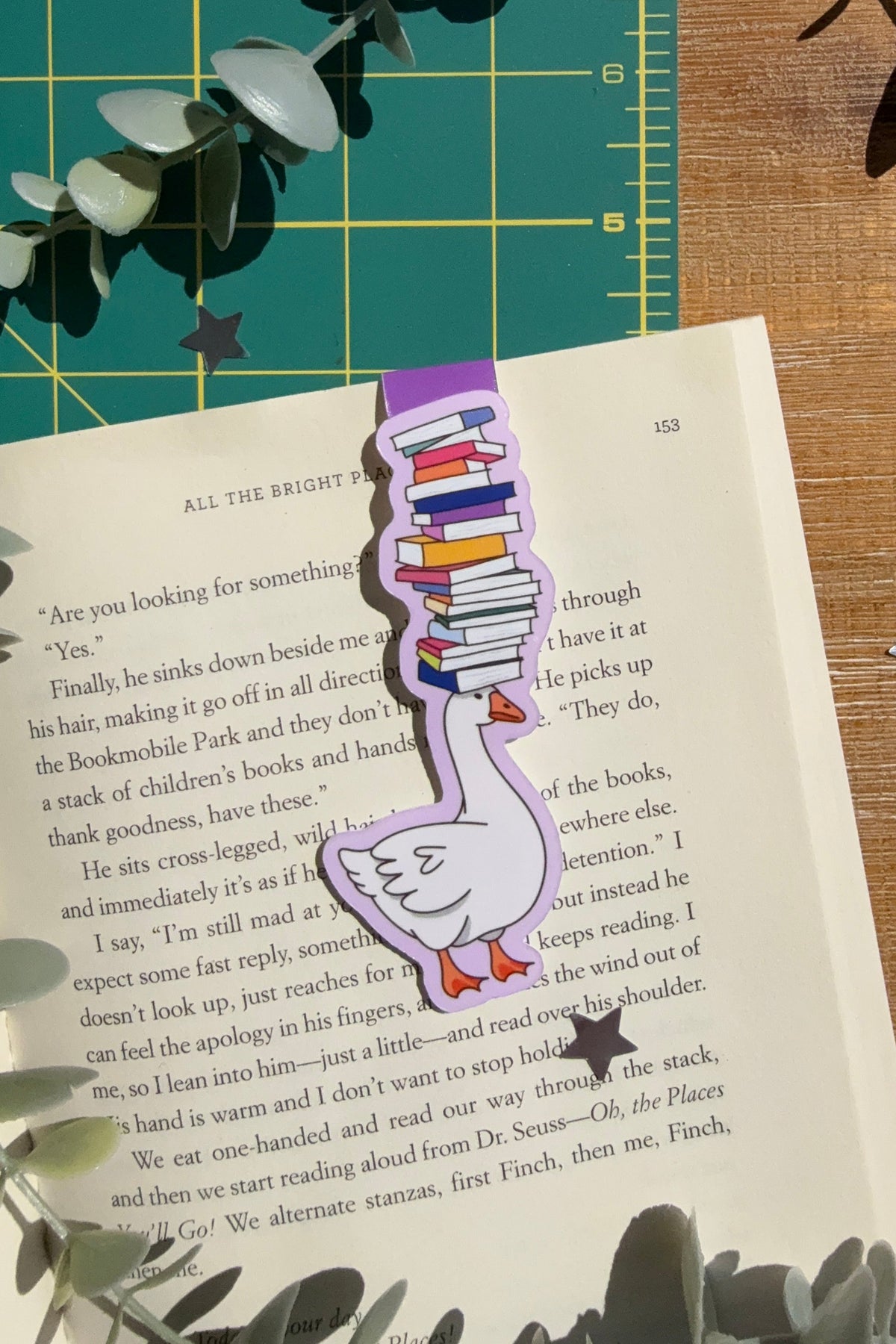 Magnet bookmark of a goose balancing a stack of books on its head on an open book with a green cutting mat and wood desk in the background.