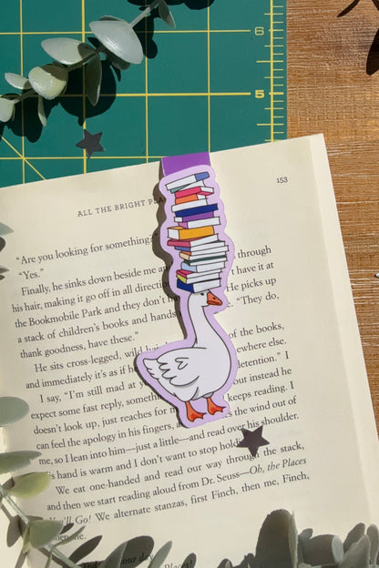 Magnet bookmark of a goose balancing a stack of books on its head on an open book with a green cutting mat and wood desk in the background.