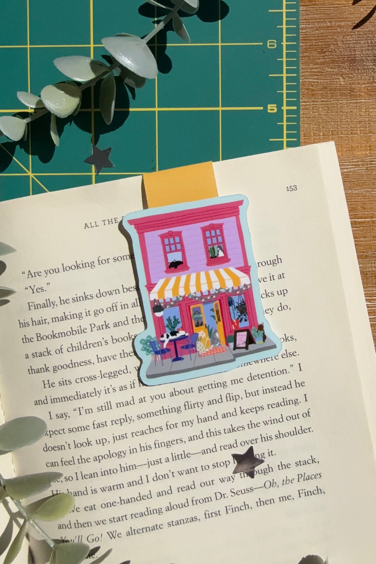 A small illustrated bookmark shaped like a pink storefront with a yellow awning and blue sky is placed on an open book, surrounded by eucalyptus leaves on a wooden surface with a green cutting mat in the background.