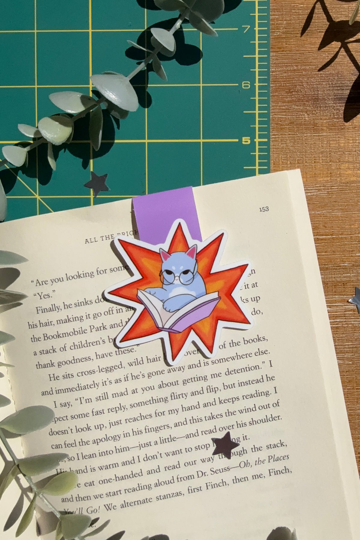 A cartoon-style book bookmark featuring a blue cat inside a red starburst shape, placed on an open book on a wooden surface with eucalyptus leaves and a green cutting mat in the background.