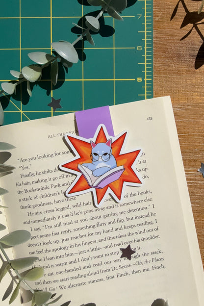 A cartoon-style book bookmark featuring a blue cat inside a red starburst shape, placed on an open book on a wooden surface with eucalyptus leaves and a green cutting mat in the background.