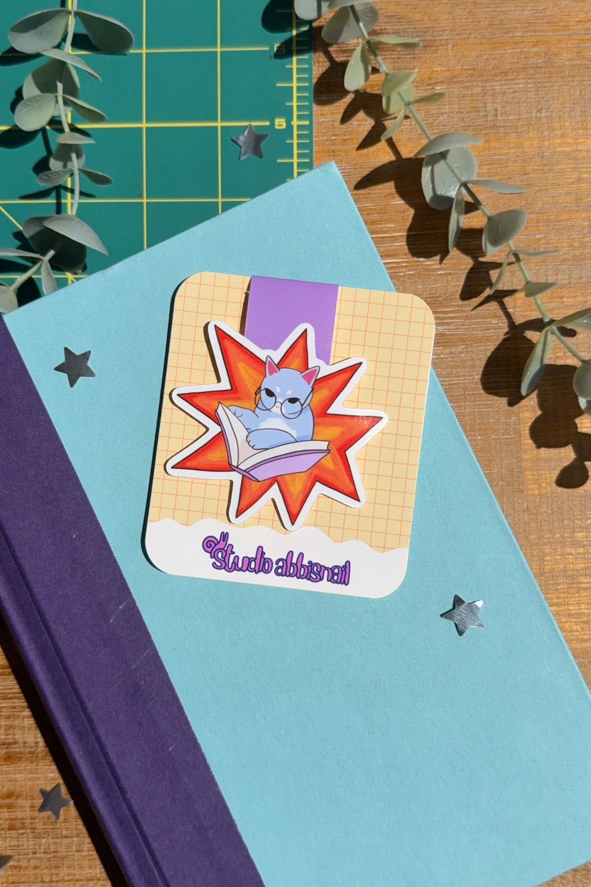 A magnet bookmark of a blue cat with glasses reading in an orange explosion, sitting on a blue book with a green cutting mat and wood desk in the background.