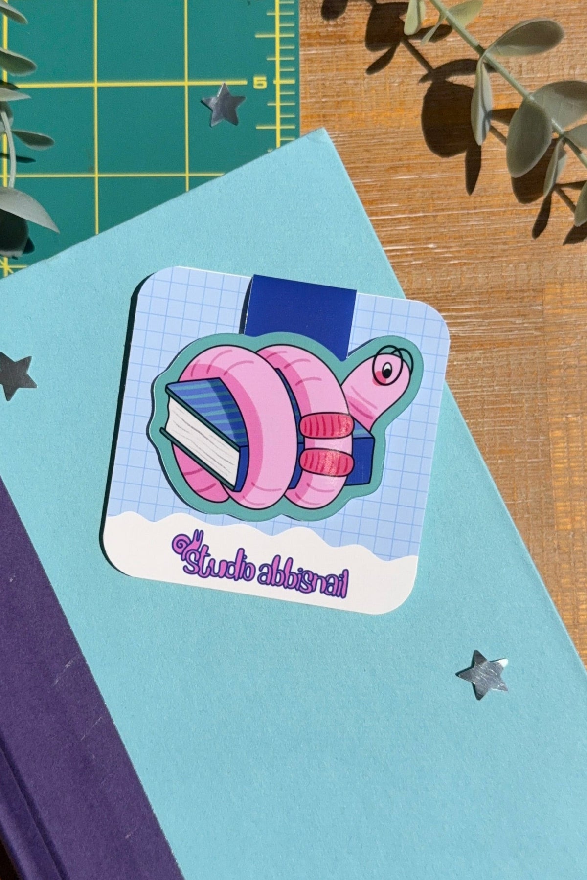 A square sticker featuring a pink and blue cartoon worm with glasses wrapped around a book, placed on a light blue book, styled on a wooden desk with a green cutting mat and eucalyptus leaves.