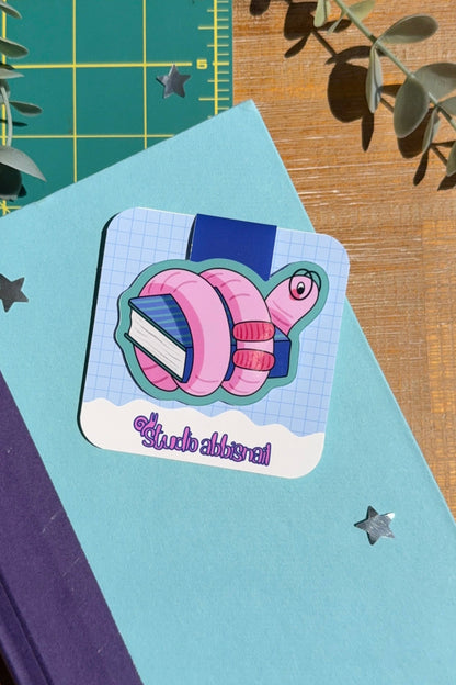 A square sticker featuring a pink and blue cartoon worm with glasses wrapped around a book, placed on a light blue book, styled on a wooden desk with a green cutting mat and eucalyptus leaves.