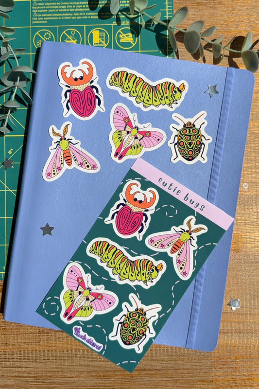 'Cutie bugs' sticker sheet with individual stickers on blue notebook with a wood and green cutting mat background.