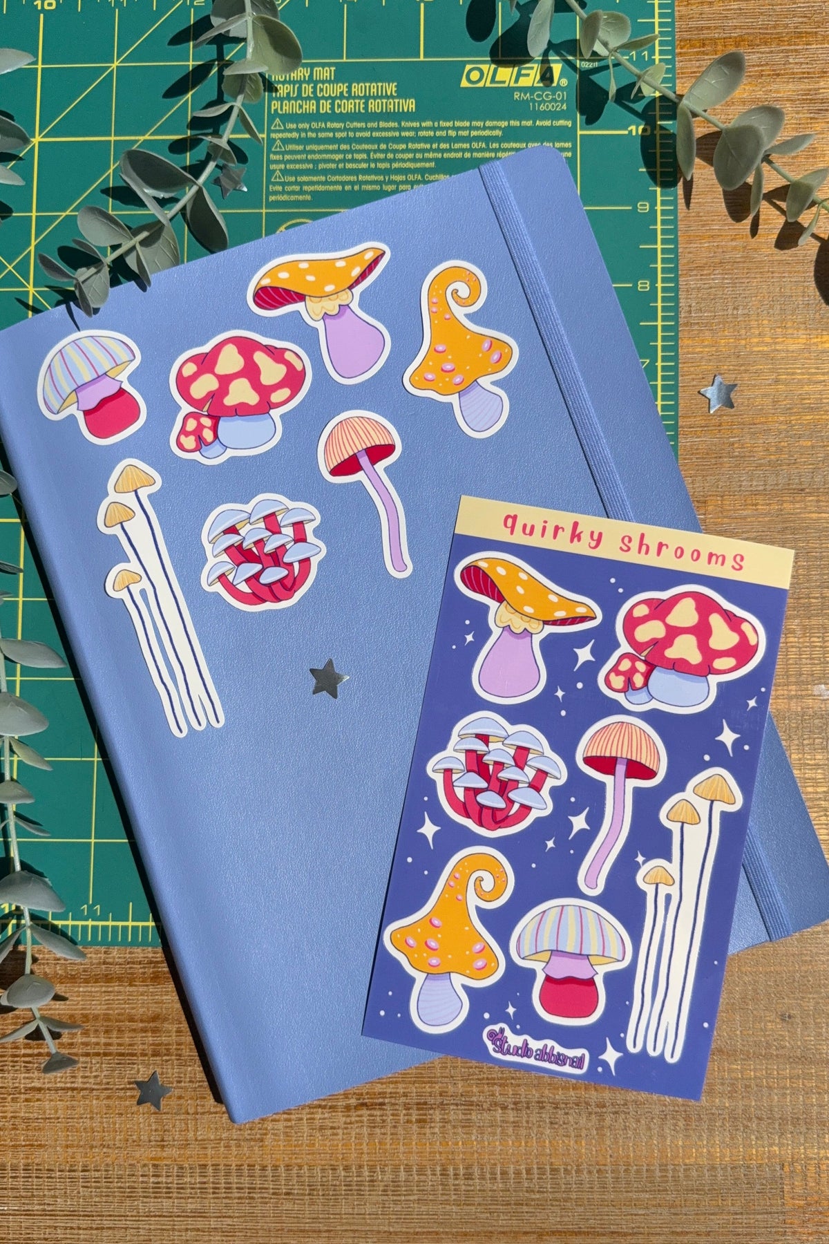 A blue notebook and a matching sticker sheet featuring whimsical mushroom illustrations, styled on a wooden surface with a green cutting mat and eucalyptus leaves.