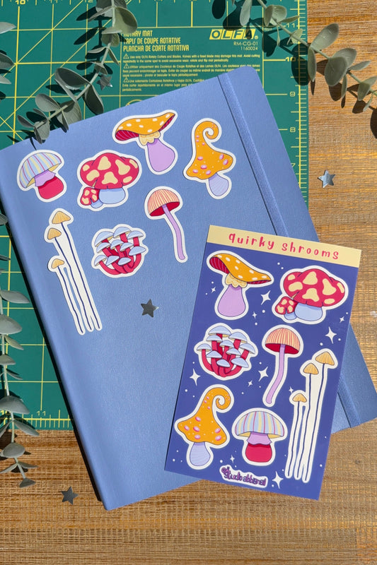 A blue notebook and a matching sticker sheet featuring whimsical mushroom illustrations, styled on a wooden surface with a green cutting mat and eucalyptus leaves.