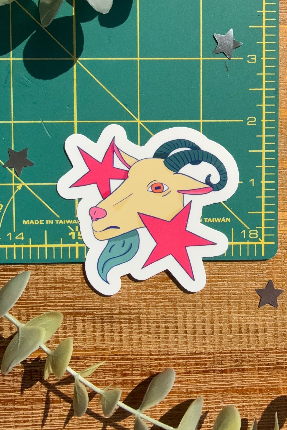 A die-cut sticker featuring a stylized cartoon ram head surrounded by pink stars, placed on a green cutting mat over a wooden surface with eucalyptus leaves.