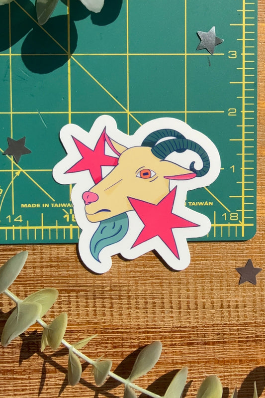 A die-cut sticker featuring a stylized cartoon ram head surrounded by pink stars, placed on a green cutting mat over a wooden surface with eucalyptus leaves.
