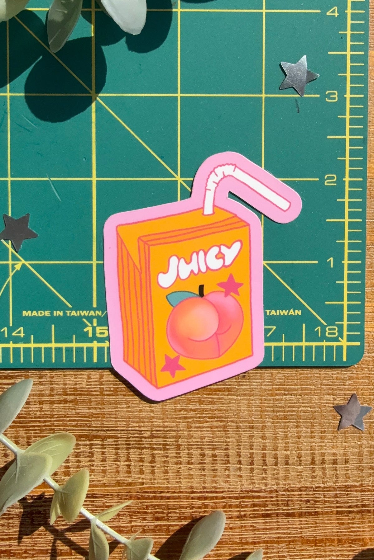 A die-cut sticker shaped like a juice box with the word 'JUICY' and a peach illustration, placed on a wooden surface with a green cutting mat and eucalyptus leaves as props.