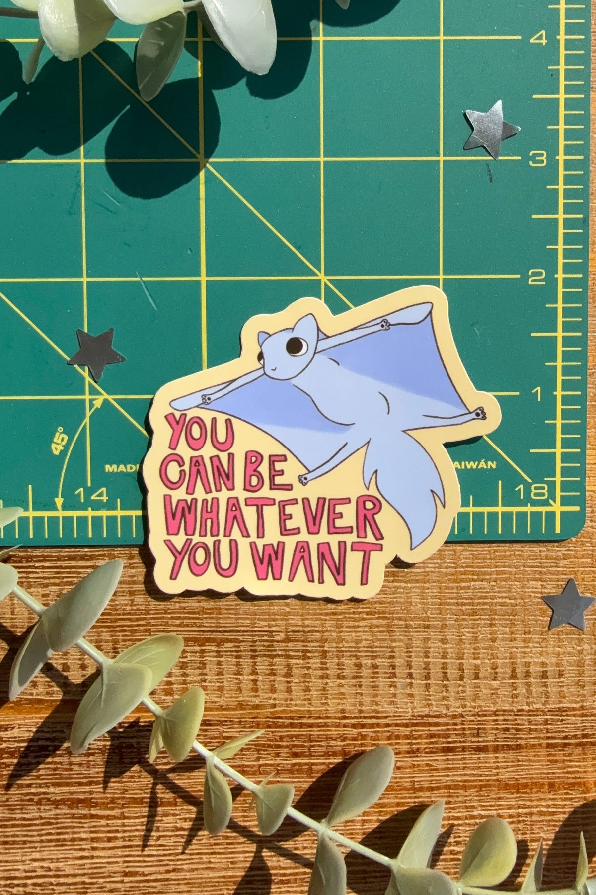 A die-cut sticker featuring a light blue flying squirrel with the text 'YOU CAN BE, WHATEVER YOU WANT' in red and yellow lettering, placed on a green cutting mat over a wooden surface with eucalyptus leaves.