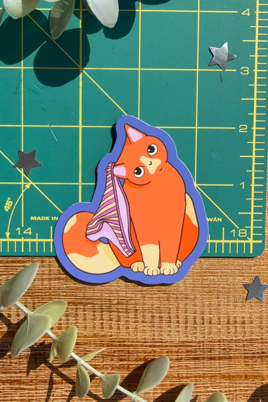 A die-cut sticker of an orange cat with a pink undies, placed on a wooden surface with a green cutting mat and eucalyptus leaves.