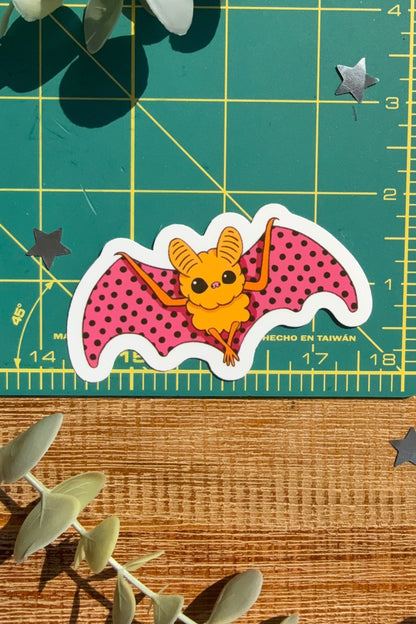 A die-cut sticker of a cute cartoon bat with pink polka-dot wings and a yellow body, displayed on a green cutting mat with eucalyptus leaves and star stickers.