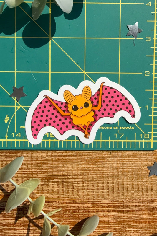 A die-cut sticker of a cute cartoon bat with pink polka-dot wings and a yellow body, displayed on a green cutting mat with eucalyptus leaves and star stickers.