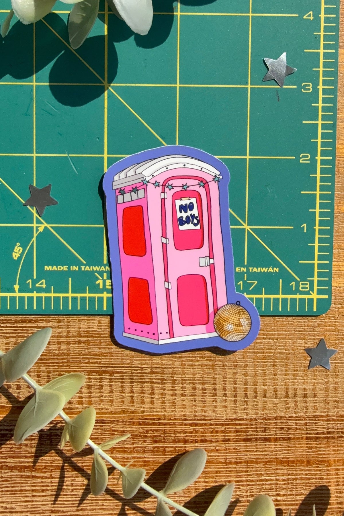 A die-cut sticker of a pink portable toilet with a purple border, placed on a wooden surface next to a green cutting mat and eucalyptus leaves.