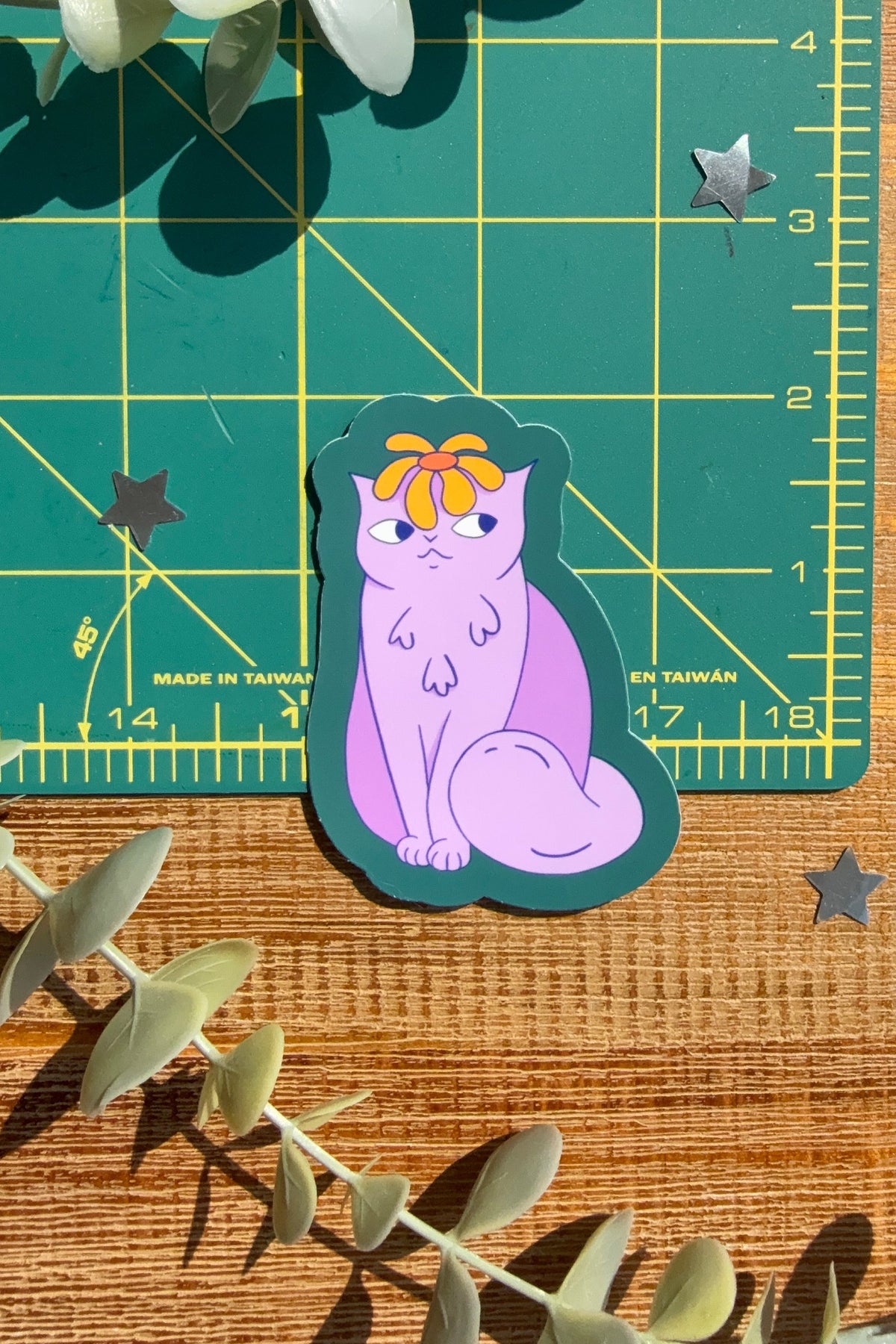 A die-cut sticker featuring a cute purple cat with a yellow flower, placed on a green cutting mat over a wooden surface with eucalyptus leaves.
