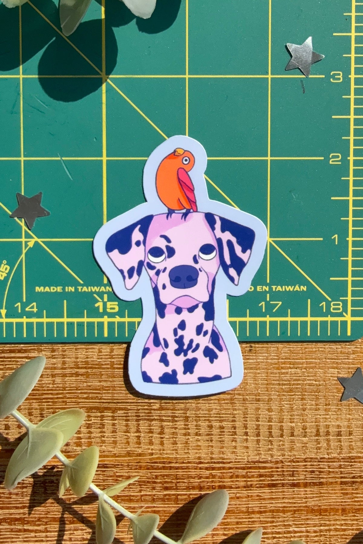 A die-cut sticker of a Dalmatian dog with an orange parrot on its head, placed on a wooden surface next to a green cutting mat and eucalyptus leaves.