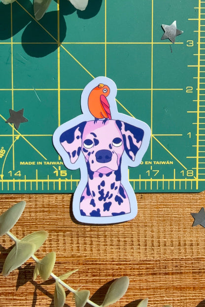 A die-cut sticker of a Dalmatian dog with an orange parrot on its head, placed on a wooden surface next to a green cutting mat and eucalyptus leaves.