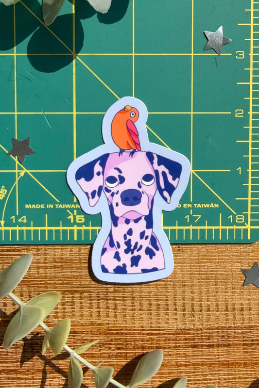 A die-cut sticker of a Dalmatian dog with an orange parrot on its head, placed on a wooden surface next to a green cutting mat and eucalyptus leaves.