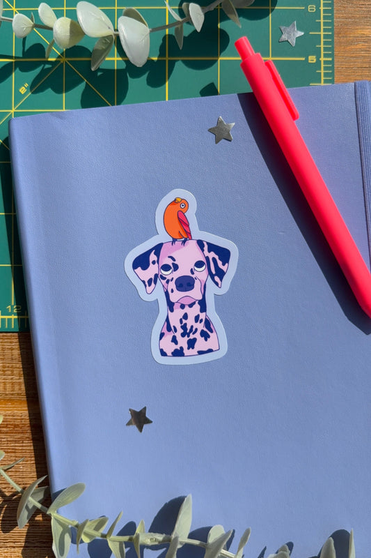 A blue notebook with a sticker of a Dalmatian dog with an orange bird on its head, styled on a green cutting mat with a red pen, eucalyptus leaves, and star stickers.