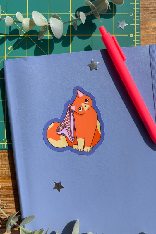 A blue notebook with a die-cut sticker of an orange cat , with pink undies styled on a green cutting mat with a red pen and eucalyptus leaves.