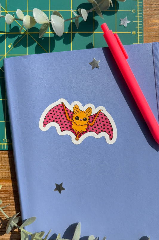A blue notebook with a cute bat sticker on the cover, styled with a pink pen, eucalyptus leaves, and a green cutting mat.