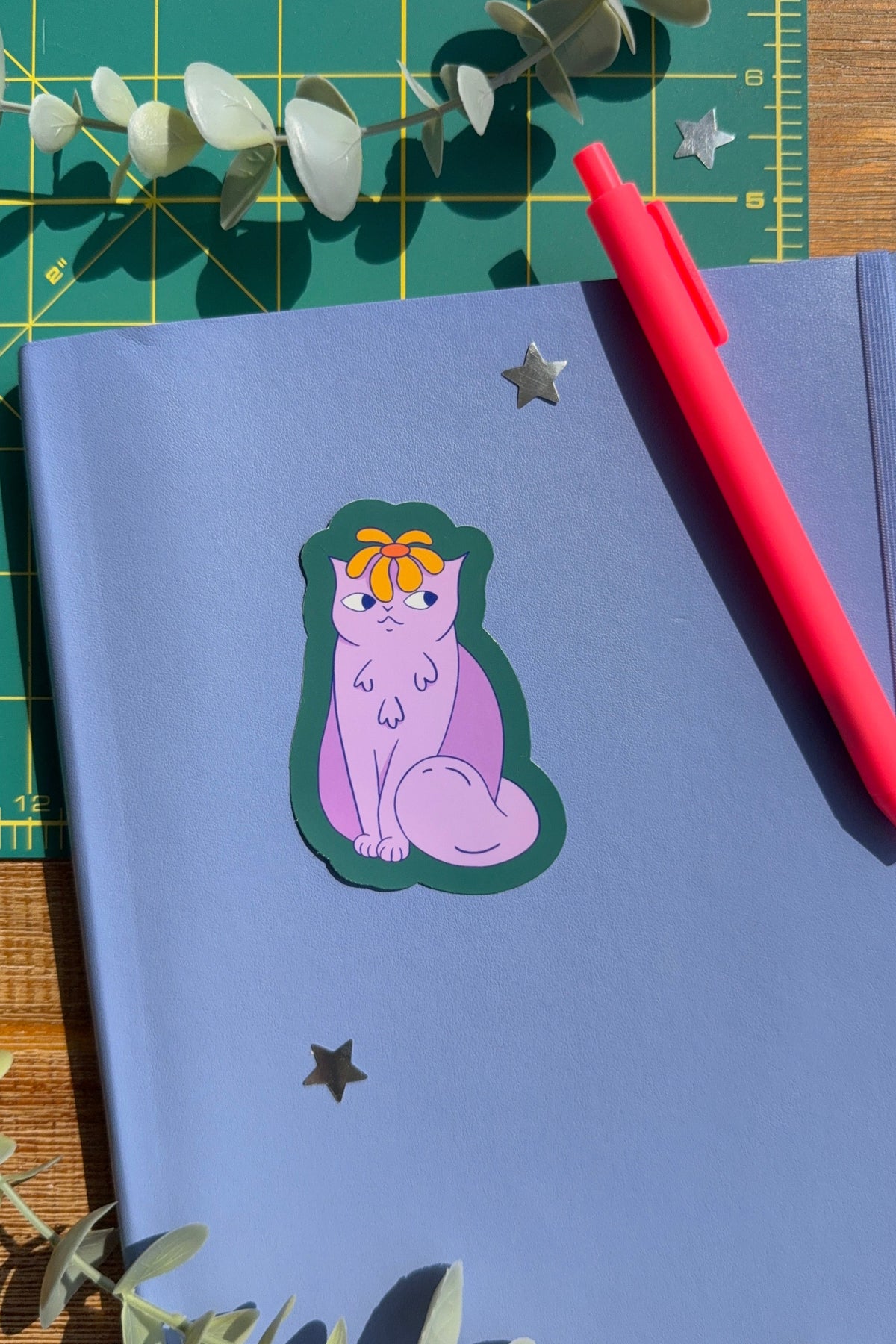 A styled flat-lay of a lavender notebook with a die-cut sticker of a purple cat wearing an orange bow, placed on a green cutting mat with eucalyptus leaves and a red pen.