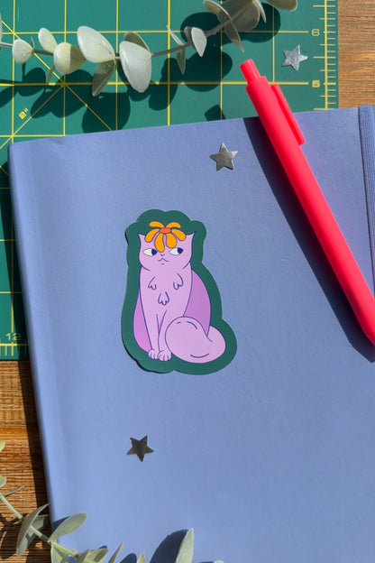 A styled flat-lay of a lavender notebook with a die-cut sticker of a purple cat wearing an orange bow, placed on a green cutting mat with eucalyptus leaves and a red pen.