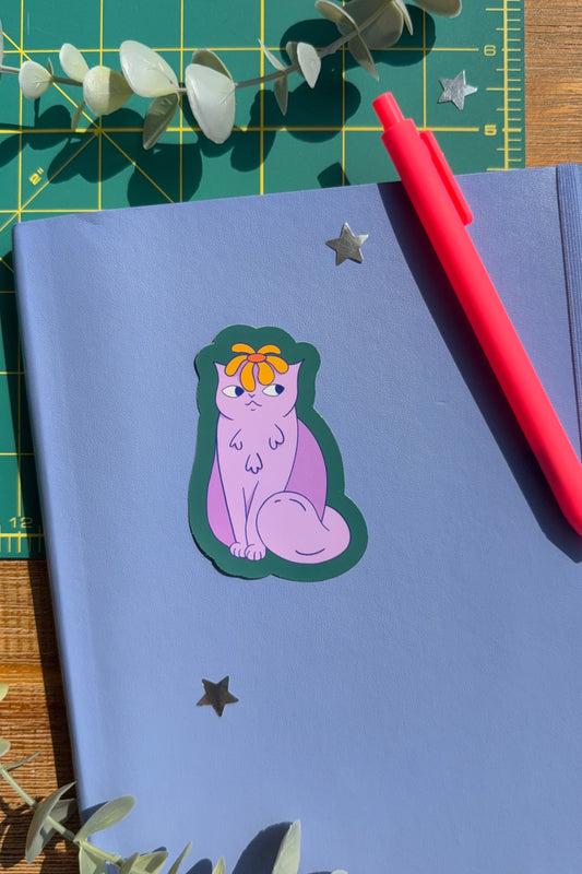A styled flat-lay of a lavender notebook with a die-cut sticker of a purple cat wearing an orange bow, placed on a green cutting mat with eucalyptus leaves and a red pen.