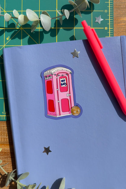 A blue notebook decorated with a pink and red porta potty sticker, a red pen, and star-shaped stickers, styled on a green cutting mat with eucalyptus leaves.