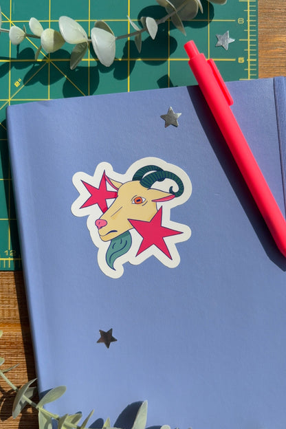 A blue notebook with a die-cut sticker of a goat head surrounded by pink stars, styled on a green cutting mat with a red pen and eucalyptus leaves.