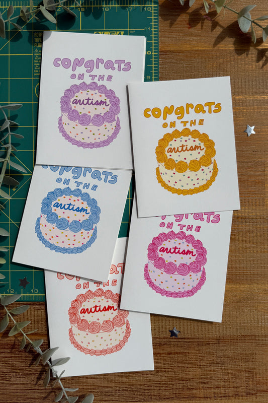 Five small greeting cards with colorful 'Congrats on the Autism' cupcake illustrations are arranged on a wooden surface with a green cutting mat and eucalyptus sprigs.