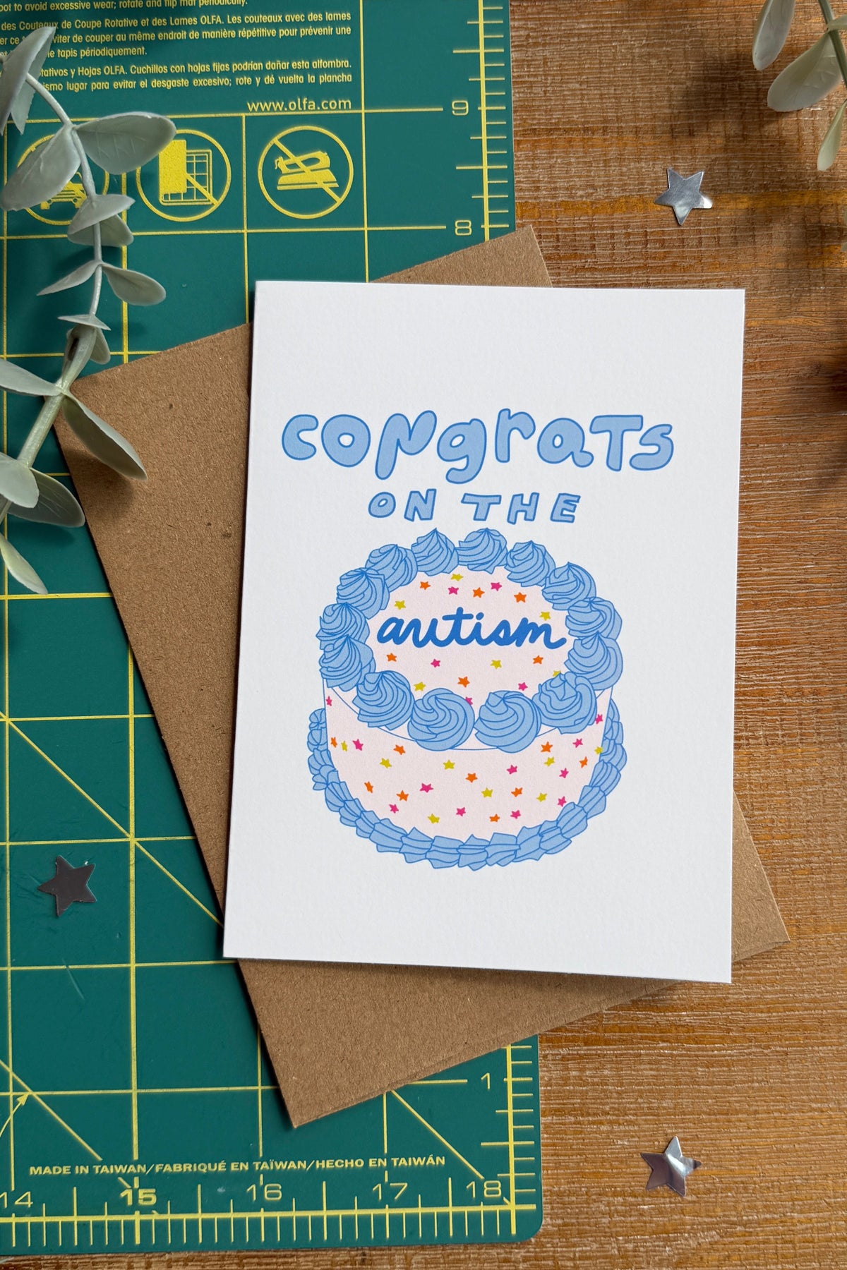 A white greeting card with a blue and pink illustrated cake and the words 'congrats on the autism' lies on top of a brown kraft envelope, styled on a wooden surface with a green cutting mat and eucalyptus branches.