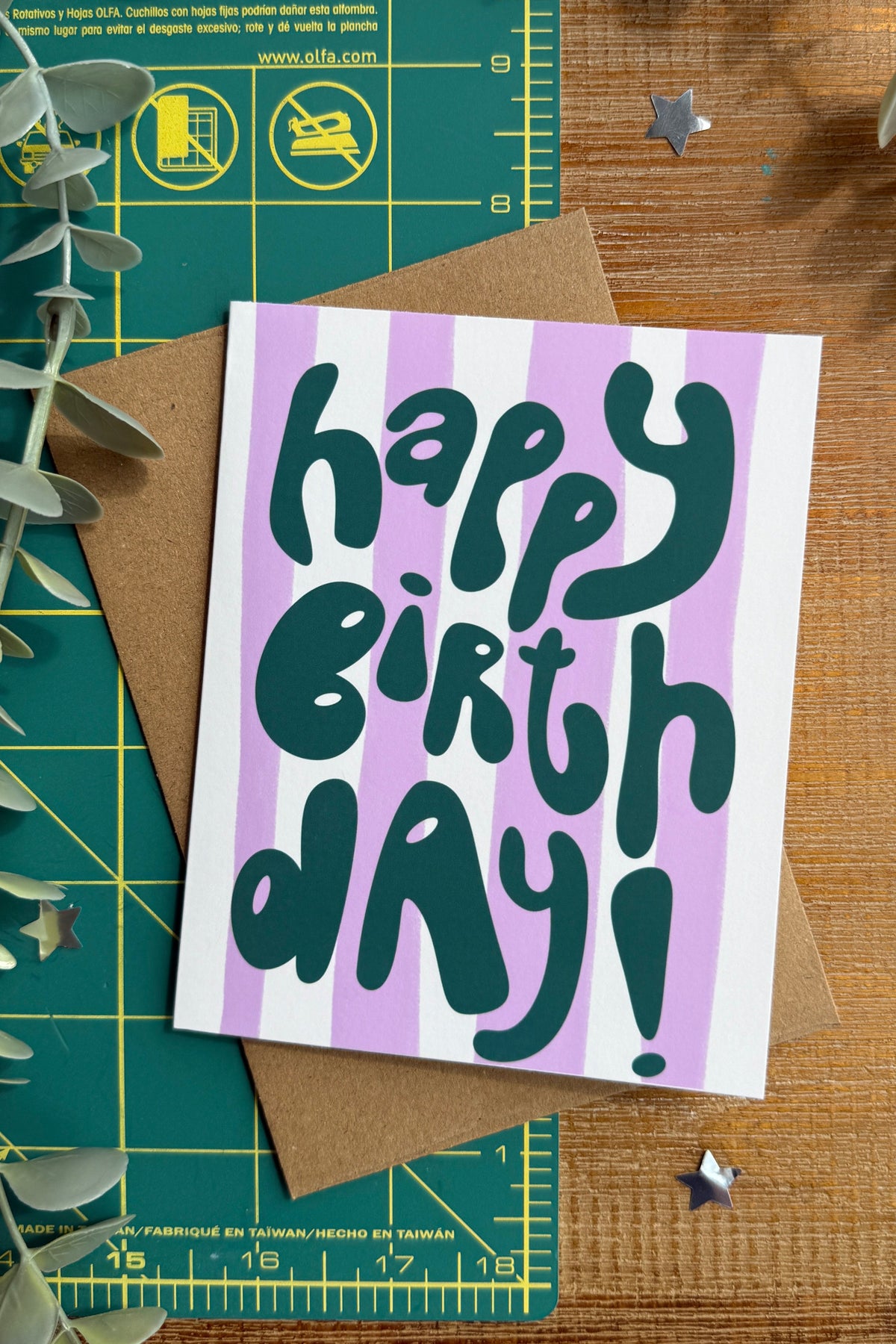 A square birthday card with the words 'happy birthday!' in dark teal lettering over lavender and white vertical stripes, placed on top of a brown kraft envelope on a wooden surface with eucalyptus leaves and a green cutting mat.