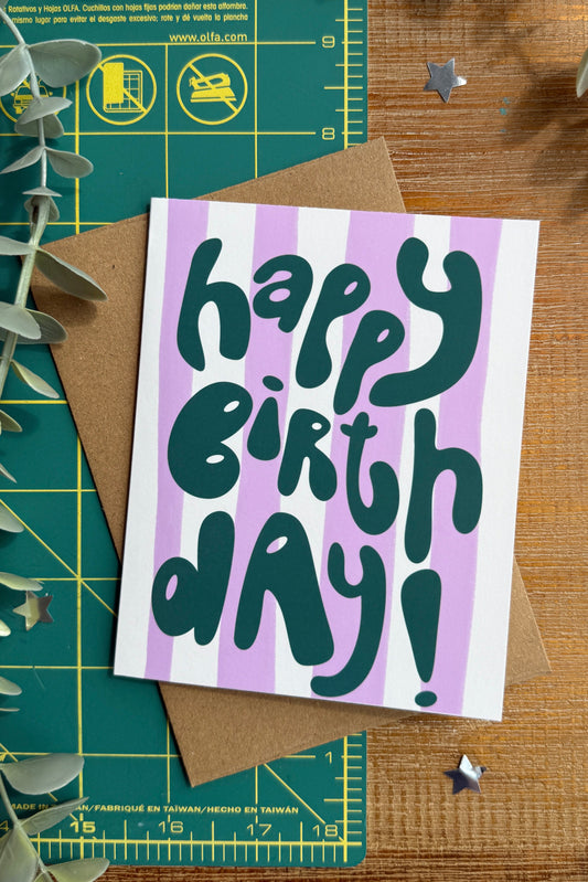 A square birthday card with the words 'happy birthday!' in dark teal lettering over lavender and white vertical stripes, placed on top of a brown kraft envelope on a wooden surface with eucalyptus leaves and a green cutting mat.
