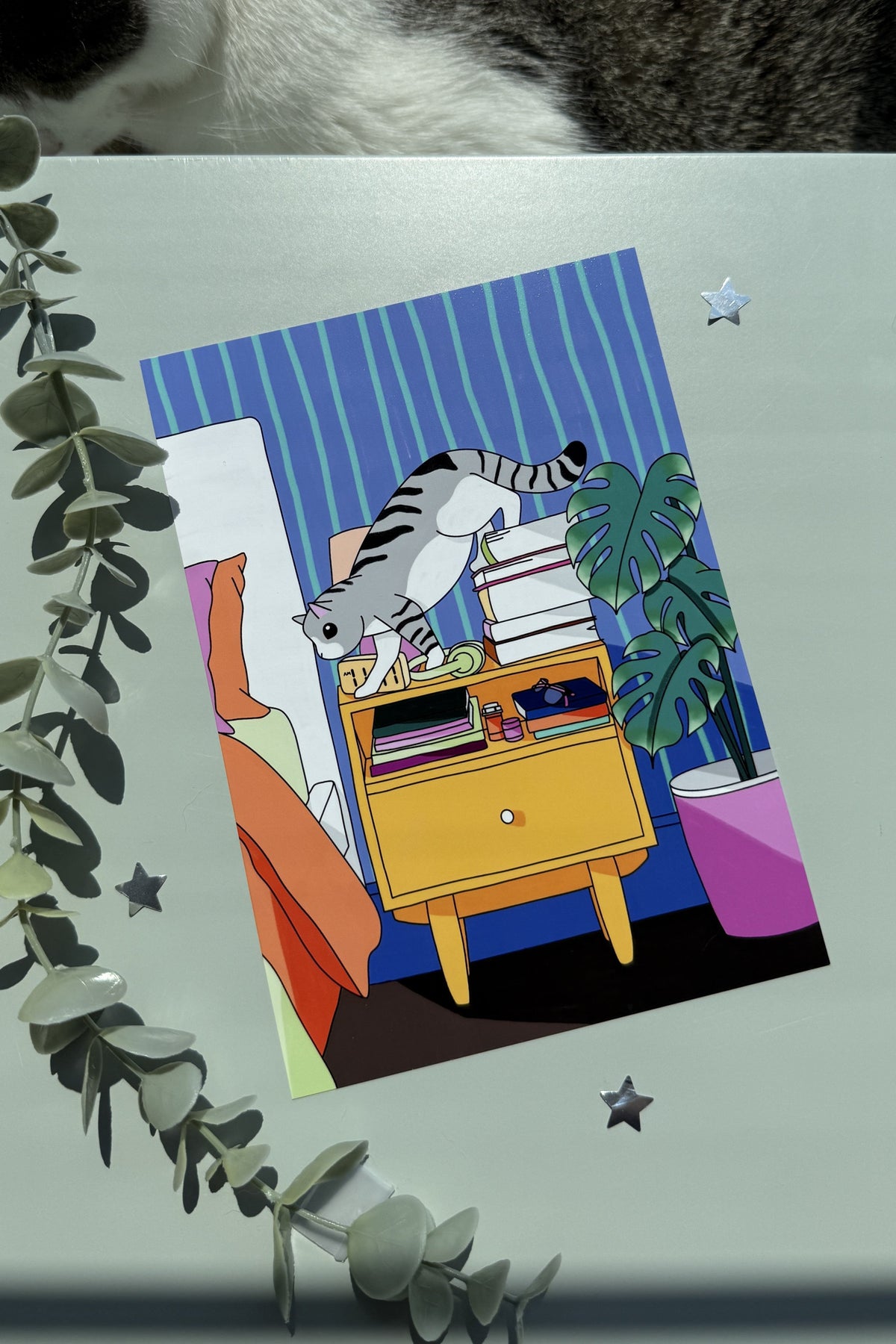 A square art print featuring a colorful cartoon illustration of a gray tabby cat climbing on books next to a yellow cabinet and a green plant, displayed on a light gray surface with decorative eucalyptus leaves and star stickers. A real cat is partially visible at the top edge of the frame.