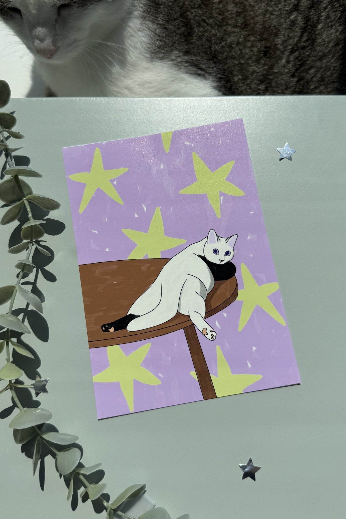 A square art print featuring a white cat lounging on a wooden table against a lavender background with yellow stars, styled on a tabletop with a real cat partially visible at the top and greenery at the side.
