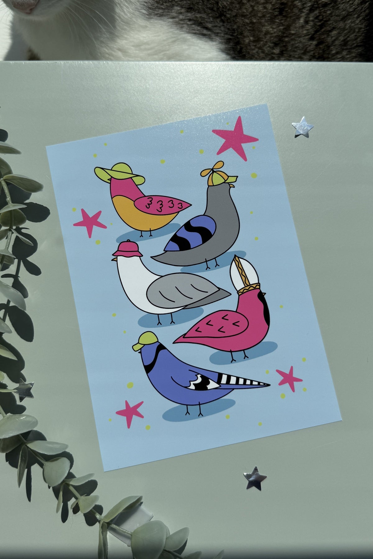 A square art print featuring a whimsical illustration of five birds in pink, blue, yellow, and gray on a light blue background, styled on a gray surface with a cat partially visible at the top and greenery at the side.