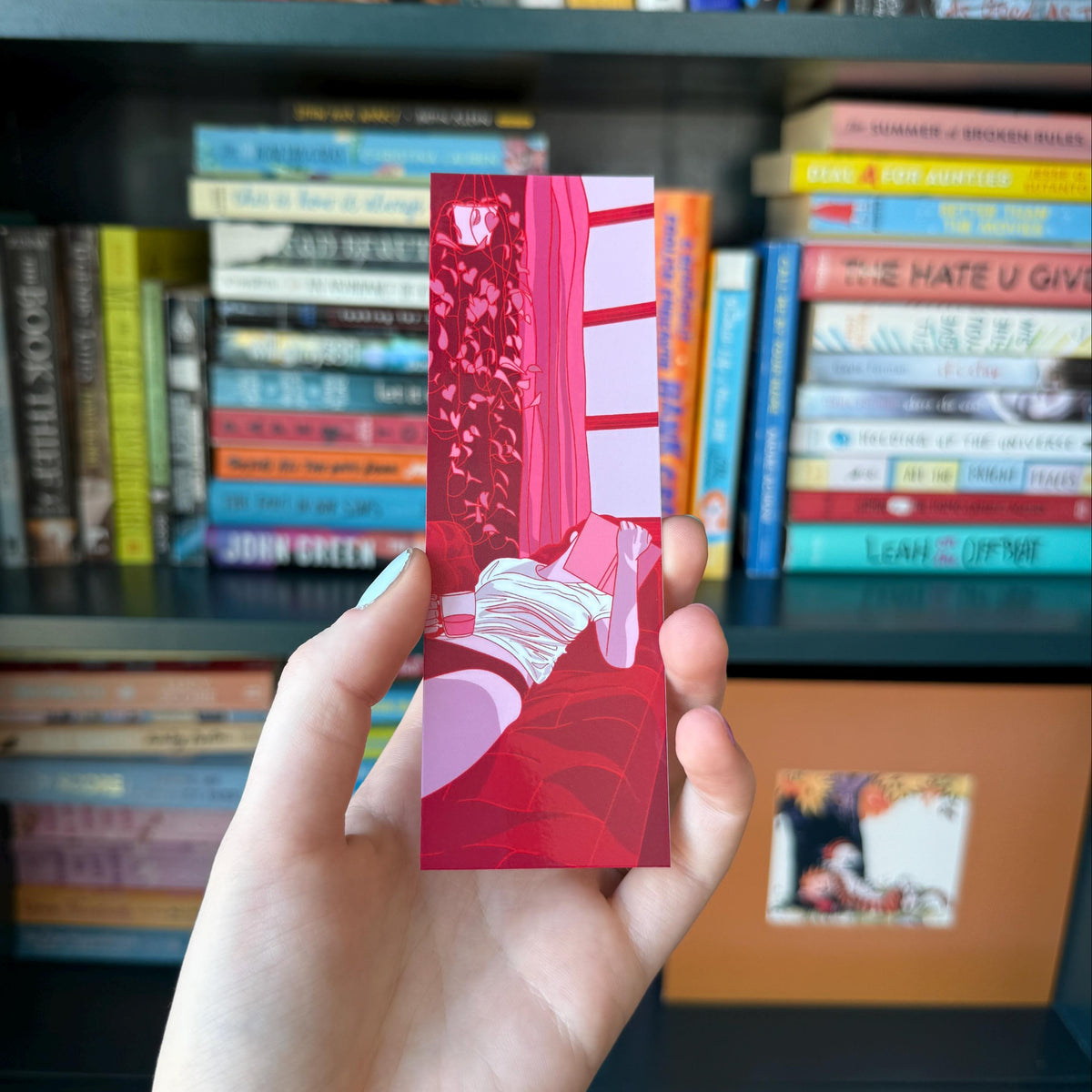 A hand holds a vertical art print featuring a stylized illustration of a pink and red room with a window, set against a blurred background of bookshelves filled with books.